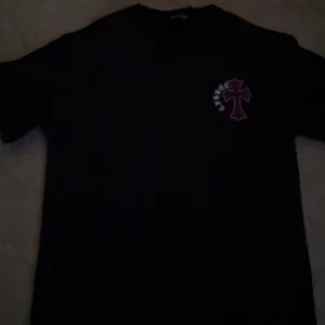 T-shirt Chrome Hearts - Svart t-shirt från Chrome Hearts med stort lila korsmotiv och vit text på ryggen samt mindre tryck på bröstet. Klassisk passform och rund hals. Perfekt för dig som gillar streetwear och vill sticka ut med en unik design.