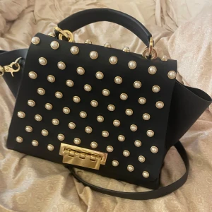 Zac Zac Posen handväska med pärldetaljer - Säljer denna handväskan från Zac Zac Posen i marinblått skinn med guldfärgade metalldetaljer och pärlliknande nitar över hela framsidan. Väskan har både handtag och axelrem, samt en unik låsdetalj framtill. Jättebra skick och aldrig använd. Den har lite mini små repor på guld knäppet men annars inte en skråma. Pris går att diskutera 🪽💗