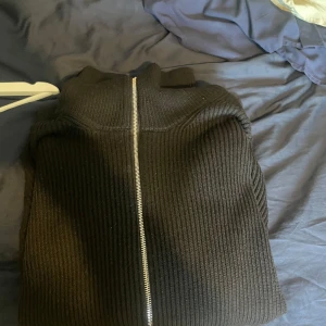 Svart ribbad kofta med dragkedja - Säljer en svart ribbad kofta med hel dragkedja framtill. Koftan har en hög krage och är stickad i ett tjockare material som känns både mjukt och varmt. Perfekt för dig som gillar en clean och stilren look med lite edge.