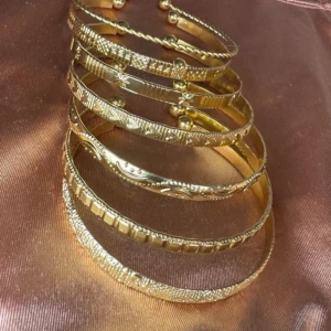 Set med 7 Guldiga armband med mönster - Säljer ett set med 7 guldiga armband i olika mönster och strukturer. Armbanden har detaljer som vridna och graverade ytor, vilket ger en lyxig och unik känsla. Perfekt för att ge din stil lite extra bling och personlighet.