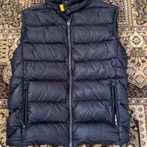  Parajumpers väst - Snygg svart dunväst från Parajumpers, modell SHEEN. Västen har quiltad design, två dragkedjeförsedda fickor och hög krage med gul detalj. Insidan är ljusgrå och har flera etiketter med coola detaljer. Perfekt att slänga på över en hoodie.