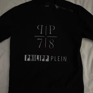 Svart Philipp Plein sweatshirt med dragkedjor !ÄKTA! - Svart sweatshirt från Philipp Plein med silverfärgad logga framtill och metallmärke i nacken. Tröjan har långa ärmar, coola dragkedjor på axel och sida samt ribbade muddar. Snygg och stilren med tydlig streetkänsla.