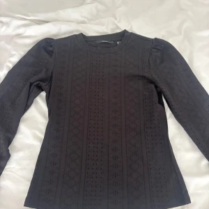 Svart blus med hålmönster Vero Moda - Supersnygg svart blus från Vero Moda med lång ärm och diskret hålmönster i geometriska ränder. Puffade axlar ger en trendig siluett och materialet känns mjukt mot huden. Perfekt för dig som gillar stilrena detaljer och vill sticka ut lite extra.
