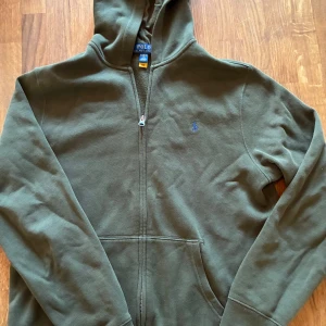 Polo Ralph Lauren hoodie - Olivgrön hoodie från Polo Ralph Lauren i storlek XL barn, sitter som en xs. Klassisk design med dragkedja, huva och känguruficka. Broderad blå logga på bröstet. Tillverkad i mjuk bomullsblandning med polyester för extra komfort. Perfekt för chill dagar och streetstyle.