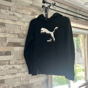 Svart hoodie från Puma med logga - Svart hoodie från Puma i storlek M med stor vit Puma-logga på bröstet. Tröjan har huva, magficka och långa ärmar. Materialet är mjukt och känns som bomull med insida i fleece. Perfekt för chill eller träning.