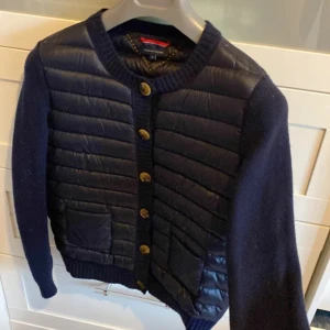 Mörkblå quiltad cardigan från Tommy Hilfiger - Snygg mörkblå quiltad cardigan från Tommy Hilfiger med quiltad framsida, två fickor och stora knappar. Västen har stickade ärmar och ribbade muddar. Materialmix av nylon, lammull och dun/fjäderfyllning. Perfekt för lager-på-lager och en clean look.Liknar cardigansen från Moncler!
