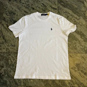 Vit t-shirt från Polo Ralph Lauren - Vit t-shirt från Polo Ralph Lauren med klassisk rund hals och korta ärmar. T-shirten har en liten broderad logga i marinblått på bröstet och är gjord i mjuk bomull. Enkel och stilren design som funkar till allt.