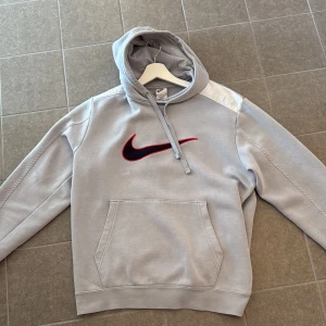 Grå Nike hoodie med svart logga - Säljer en grå hoodie från Nike med stor svart och röd logga på bröstet. Tröjan har huva med snörning, en stor magficka och långa ärmar. Perfekt för chill dagar eller när du vill ha en sportig look. Kvitto finns. Från stadium. Storlek M men passar som S.