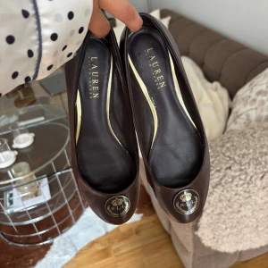 Mörkbruna ballerinaskor i skinn, Ralph Lauren - Unika vintage Ralph Lauren i skinn. Rund tå och guldfärgad RL-logga framtill. Klassisk och stilren modell med platt sula. Lite slitna baktill men annars bra skick (sista bilden).