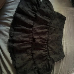 Svart volangkjol med broderade blommor - Supersöt svart kortkjol med volanger och broderade blommor. Kjolen har spetsdetaljer längs kanterna och ett broderat blommönster över hela tyget. Perfekt att styla med en enkel topp och boots.