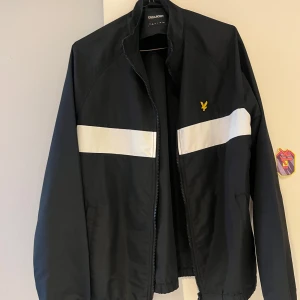 Svart vindjacka från Lyle & Scott - Svart vindjacka från Lyle & Scott med vit horisontell rand över bröstet och gul logga på vänster sida. Jackan har dragkedja framtill, ståkrage och elastisk nederkant. Perfekt för dig som gillar sportig och clean stil.