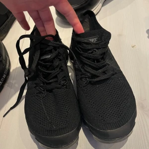 Svarta Nike Vapormax sneakers - Säljer ett par helsvarta Nike Vapormax sneakers med stickad ovandel och tydlig logga på hälen. Skorna har snörning och den ikoniska bubbliga sulan som ger en futuristisk vibe. Perfekta för dig som gillar streetstyle och vill ha något bekvämt och snyggt.