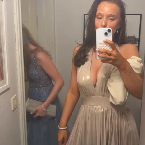 Champagnefärgad långklänning med v-ringning - Säljer en elegant champagnefärgad långklänning med djup v-ringning och bred midja. Klänningen har plisserad kjol och är ärmlös, vilket ger en lyxig och flowy känsla. Perfekt för bal eller fest. Materialet har en glansig finish som fångar ljuset snyggt.