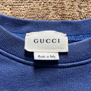 Blå tröja från Gucci - Snygg blå sweatshirt från Gucci med klassisk logga tryckt framtill i mörkare blått. Tröjan har rund halsringning och långa ärmar. Tillverkad i mjuk bomull som känns skön mot huden. Perfekt för dig som gillar stilrena designer och vill sticka ut med ett exklusivt märke. Använd 1 gång bara. En riktig bra deal om du frågar mig