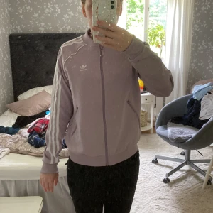 Ljuslila Adidas bomberjacka - Säljer en ljuslila Adidas bomberjacka med klassiska vita ränder längs ärmarna och dragkedja framtill. Jackan har ribbade muddar, två fickor med dragkedja och Adidas-logga på bröstet. Perfekt för dig som vill ha en sportig och snygg look. Super skönt passform och är perfekt att ha vid träning eller till vardags 👌🏼