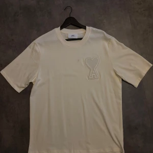 Vit t-shirt från Ami med hjärtlogo - Snygg vit t-shirt från Ami med deras ikoniska hjärta-A-logo broderad på bröstet. Klassisk passform, rund hals och tillverkad i mjuk bomull. Perfekt för en clean och stilren look.