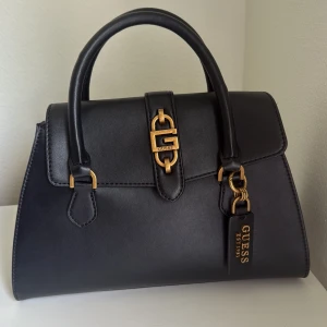 Svart handväska från Guess i skinn - Stilren svart handväska från Guess med guldfärgade metalldetaljer och logotyp framtill. Väskan har två handtag, ett rymligt fack med ljus textilfodring och innerficka med dragkedja. Klassisk och elegant design som lyfter varje outfit.