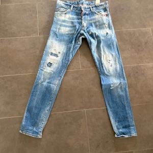 Dsquared2 ljusblå slitna jeans - Säljer ett par ljusblå jeans från Dsquared2 med coola slitningar, hål och färgstänk. Byxorna har smal passform och klassisk femficksdesign. Bakfickan har Dsquared2-logga. Perfekta för dig som gillar streetstyle och statement-plagg.