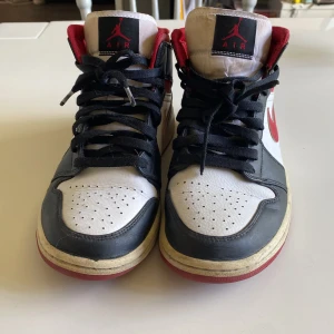 Nike Air Jordan 1 Mid svart/vit/röd - Säljer ett par Nike Air Jordan 1 Mid med klassisk färgkombination i svart, vitt och rött. Skorna har svart skinn, vit tåbox och sidor, samt röda detaljer på swoosh och häl. Hög modell med svart snörning och ikonisk Air Jordan-logga på sidan.