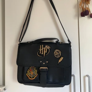Harry Potter svart axelväska - Svart axelväska i skinnimitation med justerbar rem och snygga Harry Potter-detaljer. Väskan har ett stort fack, innerficka med dragkedja och magnetlås framtill. Perfekt för dig som älskar magi och vill sticka ut. Helt oanvänd!