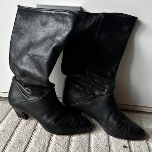 Svarta skinnstövlar med klack - Klassiska svarta stövlar i äkta skinn, tillverkade i Italien. Modellen har en rund tå och en stabil klack som ger lite höjd. Perfekta till både jeans och kjol för en snygg look. Snyggt slouchy skaft och tidlös design.