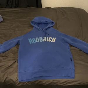 Blå HOODRICH hoodie med tryck - Säljer en blå hoodie från HOODRICH med stor logga i gradientfärger över bröstet. Tröjan har huva med snörning och långa ärmar. Perfekt för dig som gillar streetwear och vill ha en chill look.
