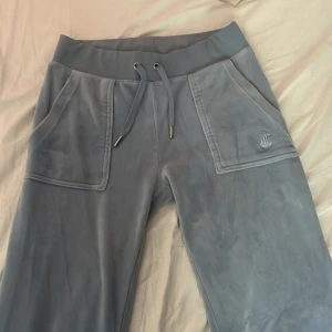 Ljusblå velourbyxor Juicy Couture XXS - Supermjuka ljusblå mjukisbyxor från Juicy Couture i velour med bred resårmidja och snörning. Byxorna har raka ben, stora fickor fram och bakfickor med knappar. Ikonisk JC-logga broderad på ena fickan. Perfekta för en chill och trendig look. Nypris 1200 kr.