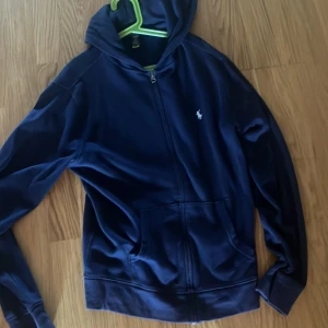 Marinblå hoodie från Polo Ralph Lauren - Snygg marinblå hoodie från Polo Ralph Lauren med dragkedja framtill och klassisk logga på bröstet. Tröjan har huva, långa ärmar och två fickor framtill. Perfekt för en chill och stilren look.