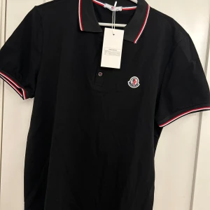 Svart pikétröja från Moncler - Svart pikétröja från Moncler med klassisk krage och knäppning framtill. Tröjan har röda och vita ränder på krage och ärmslut samt Moncler-logga broderad på bröstet. Tillverkad i bomull och har korta ärmar. Perfekt för en clean och sportig look.