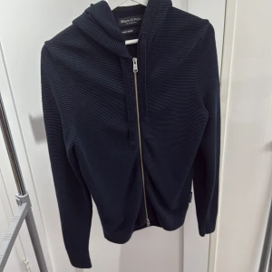 Mörkblå hoodie från Marc O'Polo - Snygg mörkblå hoodie från Marc O'Polo i ekologisk bomull. Tröjan har dragkedja framtill, huva med snörning och är ribbstickad för en modern look. Perfekt för dig som gillar stilrena och bekväma plagg. Nypris är runt 1800 