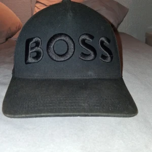 Svart keps från BOSS - Svart keps från BOSS med broderad logga framtill i svart. Justerbar rem baktill med metallspänne där BOSS är ingraverat. Klassisk böjd skärm och diskret mönstrad insida med små BOSS-loggor. Perfekt för en clean och stilren look.