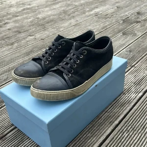 Svarta sneakers från Lanvin - Snygga svarta sneakers från Lanvin med ovandel i mocka och tåparti i skinn. Skorna har rund tå, snörning och en beige platt sula. Perfekta för dig som gillar stilrena och klassiska sneakers med exklusiv känsla.