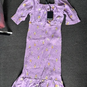 Lila blommig klänning från Pieces - Superfin lila klänning från Pieces med blommigt mönster i gult och vitt. Modellen har smockad midja, volang nertill och 3/4-ärmar. Perfekt för sommarens alla tillfällen och har en söt, feminin vibe.