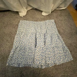 Blommig ljusblå volangkjol - Supersöt kort kjol i ljusblått med vita och svarta blommor. Kjolen har volanger och en lätt utsvängd passform som ger en luftig känsla. Perfekt för varma dagar och enkel att matcha med en enkel topp.