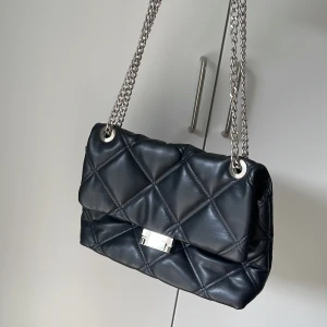 Svart quiltad axelväska med kedja - Snygg svart axelväska i quiltad skinnimitation med silverfärgad kedjerem och metallspänne. Väskan har ett klassiskt rutmönster och stilren design som ger en lyxig känsla. Perfekt storlek för det viktigaste och enkel att bära över axeln.