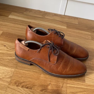 Bruna kostymskor i skinn från Strömberg - Snygga bruna skinnskor från Strömberg med klassisk design och rund tå. Skorna har mörkbruna snören och en stilren sula med kontrastsöm. Perfekta för dig som vill ha en dressad look.