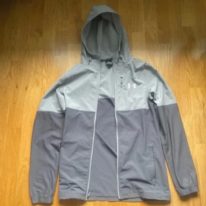 Grå Under Armour windbreaker  - Snygg tvåfärgad vindjacka från Under Armour i ljus- och mörkgrått. Jackan har huva, dragkedja hela vägen och en bröstficka med logga. Perfekt för dig som gillar sportig stil och vill ha något lätt att slänga på sig.
