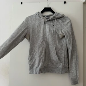 Grå hoodie från Puma med dragkedja - Säljer en grå hoodie från Puma i storlek 38. Tröjan har dragkedja framtill, två fickor och huva med snörning. Klassisk Puma-logga på bröstet och ribbade muddar vid ärmslut och nederkant. Defekt: har gula fläckar vid armhålorna. 