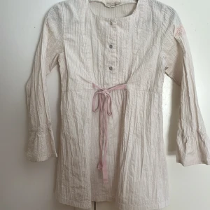 Randig blus med rosett från Tina Wodstrup - Supersöt ljusbeige och vitrandig blus från Tina Wodstrup. Blusen har långa ärmar med volangdetalj, knappar framtill och en rosa knytrosett i midjan. Broderad detalj på ärmen med TW-logga och blommor. Perfekt för dig som gillar romantisk och avslappnad stil.❤️