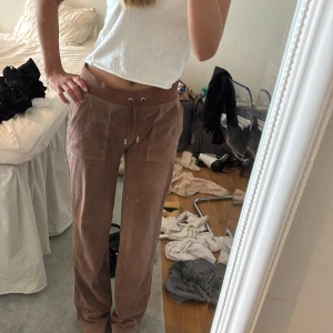 Bruna Juicy Couture mjukisbyxor - Säljer ett par bruna mjukisbyxor från Juicy Couture i mjuk velour. Byxorna har låg midja, bred resår med snörning och stora fickor både fram och bak med knappdetaljer. Perfekta för en chill och avslappnad look.