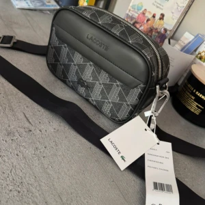 Helt ny axelväska från Lacoste  - Snygg svart axelväska från Lacoste med grått monogrammönster och justerbar rem. Väskan har silverfärgad dragkedja, ett stort huvudfack och detaljer i syntetmaterial. Perfekt för att bära det viktigaste och ge din outfit en clean vibe.Aldrig använd lapparna kvar, nypris cirka 2000kr