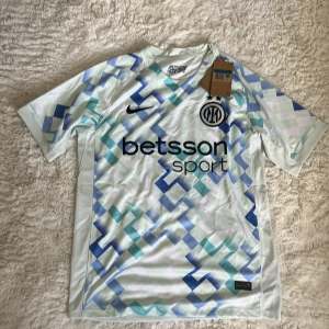 Inter Milan x Nike Dri-Fit fotbollströja  - Inter Milan x Nike Dri-Fit fotbollströja i 100% polyester. Tröjan är ljus med geometriskt mönster i blått och grönt, har korta ärmar och rund hals. Broderad Inter-logga och Nike Swoosh på bröstet samt sponsortryck. Perfekt för dig som älskar fotboll och snygg design.