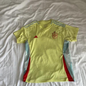 Spansk landslagströja Adidas gul - Gul fotbollströja från Adidas med Spaniens landslagsmärke på bröstet och röda detaljer. Tröjan har ljusblå ränder på axlarna, röda inslag på sidorna och ljusblå mönster längs sidorna. Tillverkad i lätt och ventilerande material, perfekt för träning eller match.
