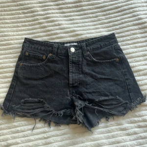 Svarta jeansshorts från Zara - Snygga svarta jeansshorts från Zara med hög midja och råa, slitna kanter. Klassisk femficksmodell med knappar framtill och fransiga detaljer nedtill för en cool, avslappnad vibe. Perfekta till sommaren och lätta att matcha med allt. Saknar en knapp, men när resten av knapparna är knäppta syns det ej!