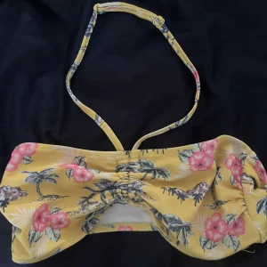Gul blommig bandeau-bikinitopp - Superfin bandeau-bikinitopp i gult med tropiskt blommönster i rosa och grönt. Toppen har smal halterneck och snygg rynkdetalj framtill. Perfekt för strandhäng och soliga dagar.