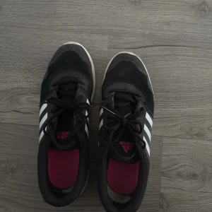 Svarta Adidas sneakers med vita ränder - Säljer ett par klassiska svarta Adidas sneakers med de ikoniska tre vita ränderna på sidorna. Skorna har snörning, platt vit sula och är tillverkade i syntet och textil. Perfekta för dig som gillar en sportig och stilren look.