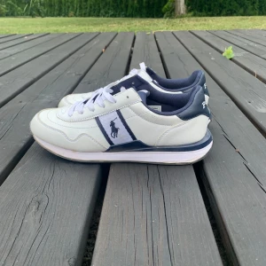 Vita sneakers från Polo Ralph Lauren - Snygga vita sneakers från Polo Ralph Lauren med mörkblå detaljer och klassisk logga på sidan. Skorna har rund tå, snörning och en platt sula. Ovandelen är i skinn och textil, med bekväm vadderad insida. Perfekta för en clean och sportig look.