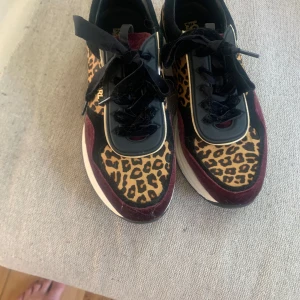 Leopardmönstrade sneakers från Karl Lagerfeld - Chunky sneakers från Karl Lagerfeld med leopardmönster, svarta och vinröda detaljer samt guldfärgade inslag. Skorna har snörning, platt sula och en cool mix av material som mocka och syntet. Perfekta för dig som vill sticka ut med din stil. 41