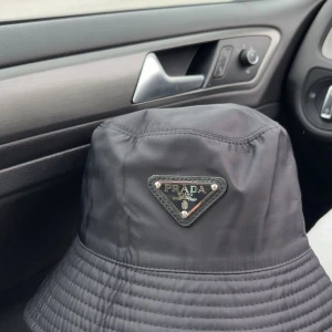 Svart bucket hat från Prada - Stilren svart bucket hat från Prada med klassisk triangellogo framtill. Hatten är tillverkad i glansig syntet och har diskreta sömmar runt brättet. Perfekt accessoar för en trendig look och enkel att matcha med olika outfits.