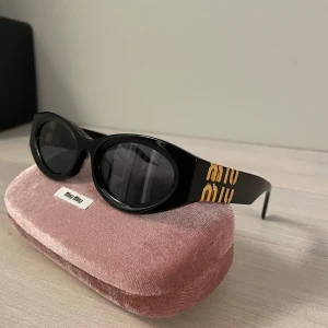 Svarta ovala solglasögon från Miu Miu - Snygga svarta solglasögon från Miu Miu med oval form och bred båge. Sidorna har guldig Miu Miu-logga som sticker ut. Glasögonen är i blank plast och kommer med ett rosa fodral i sammet med mera. Perfekt accessoar för en trendig look.